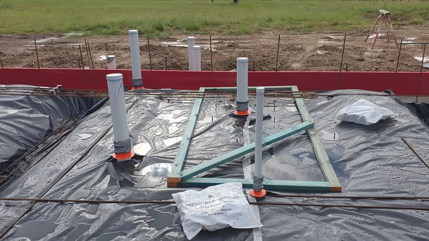 A house slab before pouring: vapour barrier down, reinforcing mesh laid, and orange collars fitted around the service penetrations